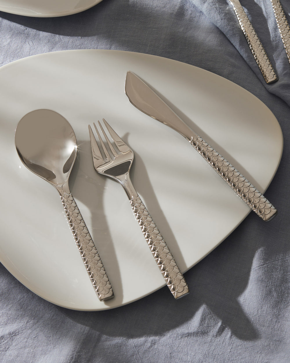 Silver flatware set by Alessi with textured fish-scale pattern handles, displayed on a white plate and blue tablecloth.