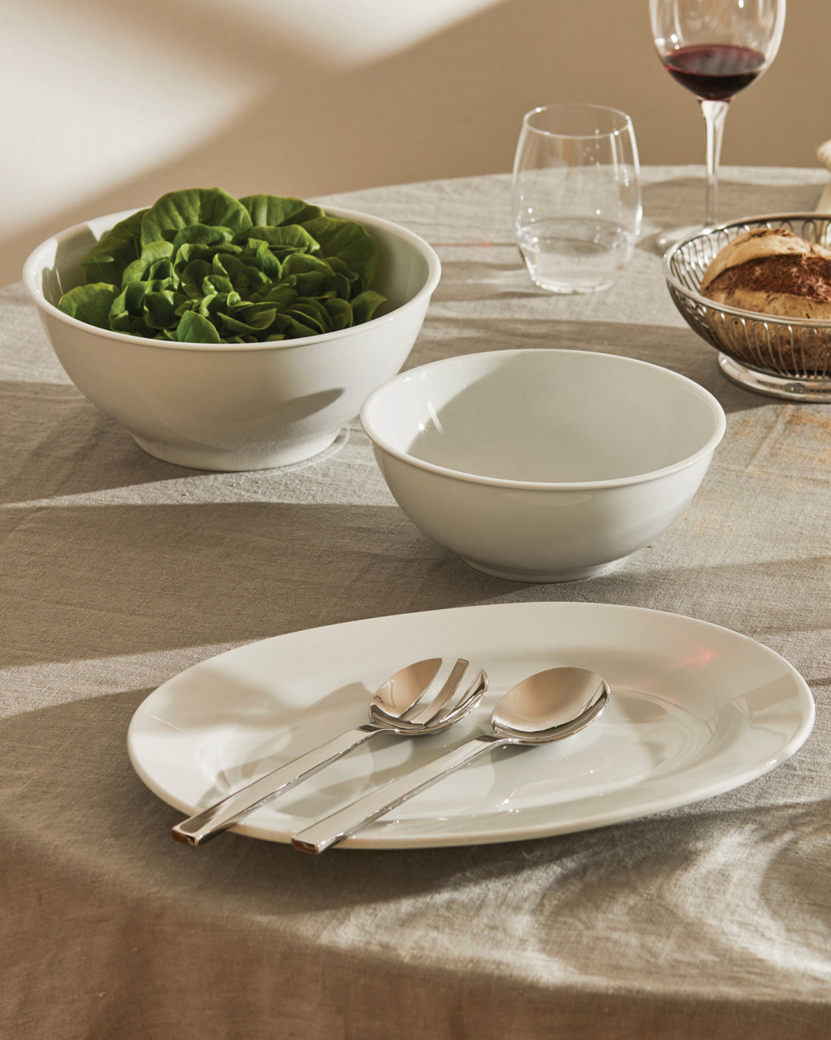 White oval plate by Alessi, styled with two bowls and salad servers on a linen tablecloth.