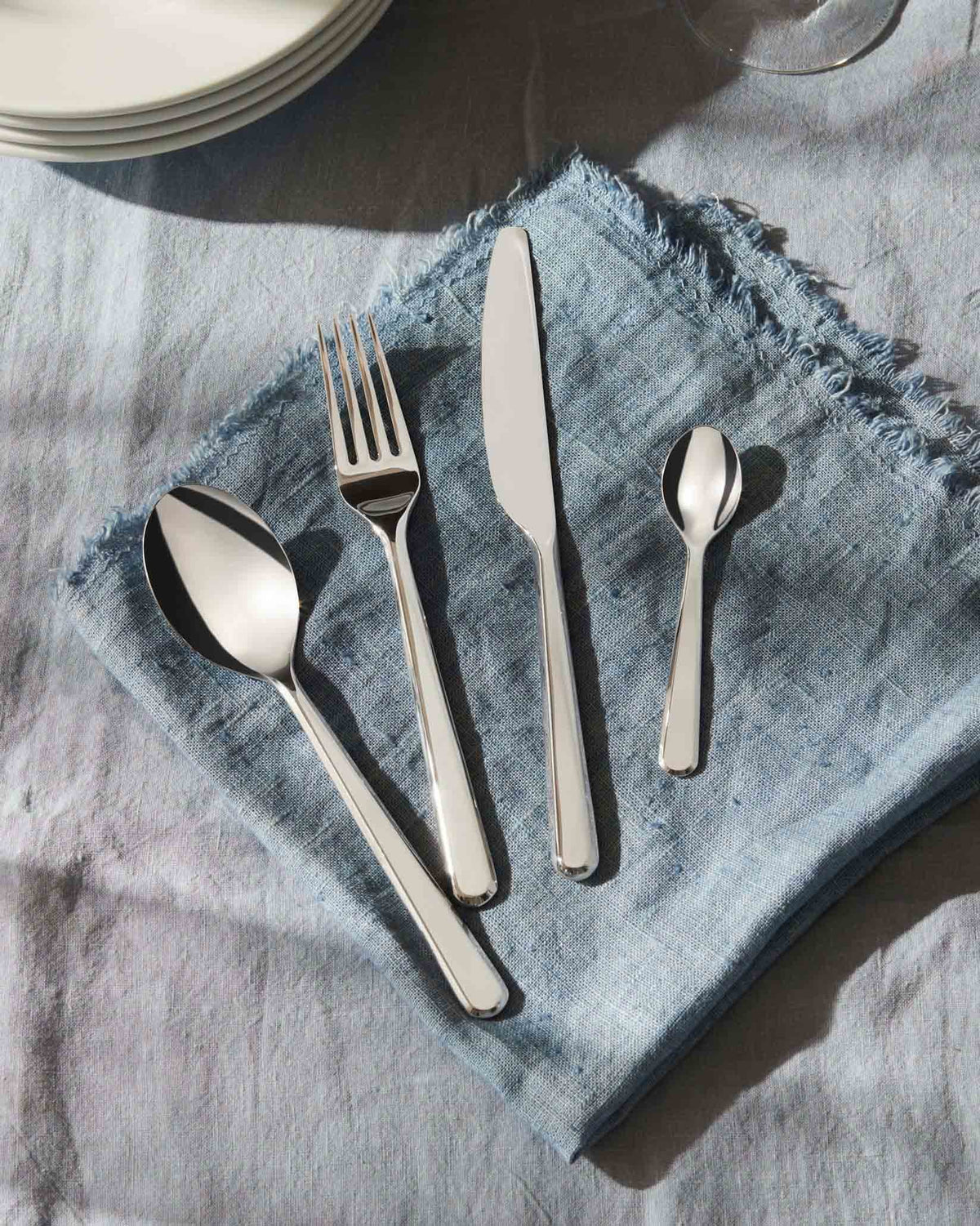 Polished silver flatware set by Alessi, with a spoon, fork, knife, and small spoon on a fringed blue linen napkin.