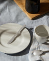 Modern dining collection by Alessi, featuring light gray dinnerware and silver cutlery on a blue linen tablecloth, with a striped napkin, wooden board, and dark jug.
