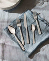 Silver flatware set by Alessi, with five polished pieces on a blue linen napkin and white plates.