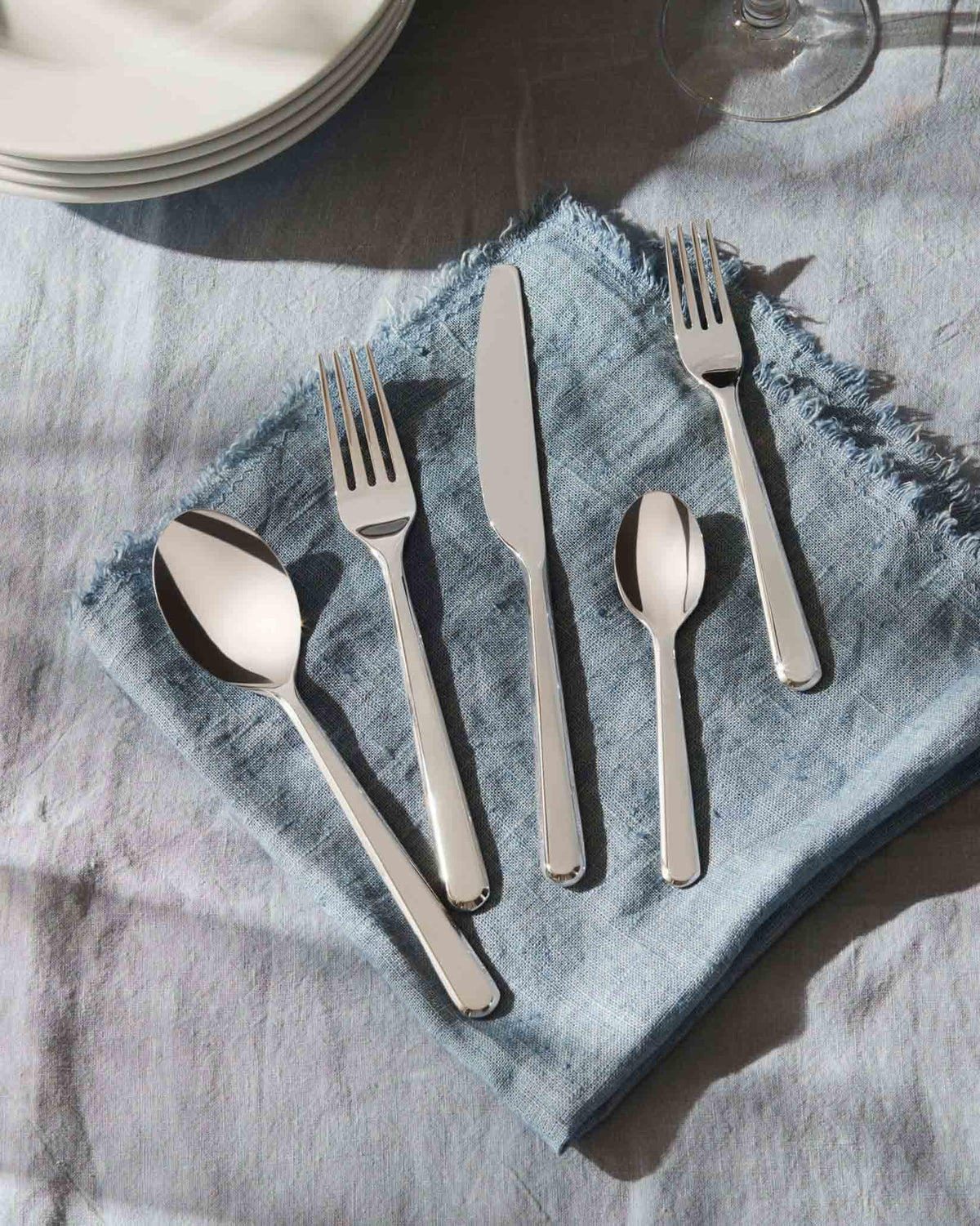Silver flatware set by Alessi, with five polished pieces on a blue linen napkin and white plates.