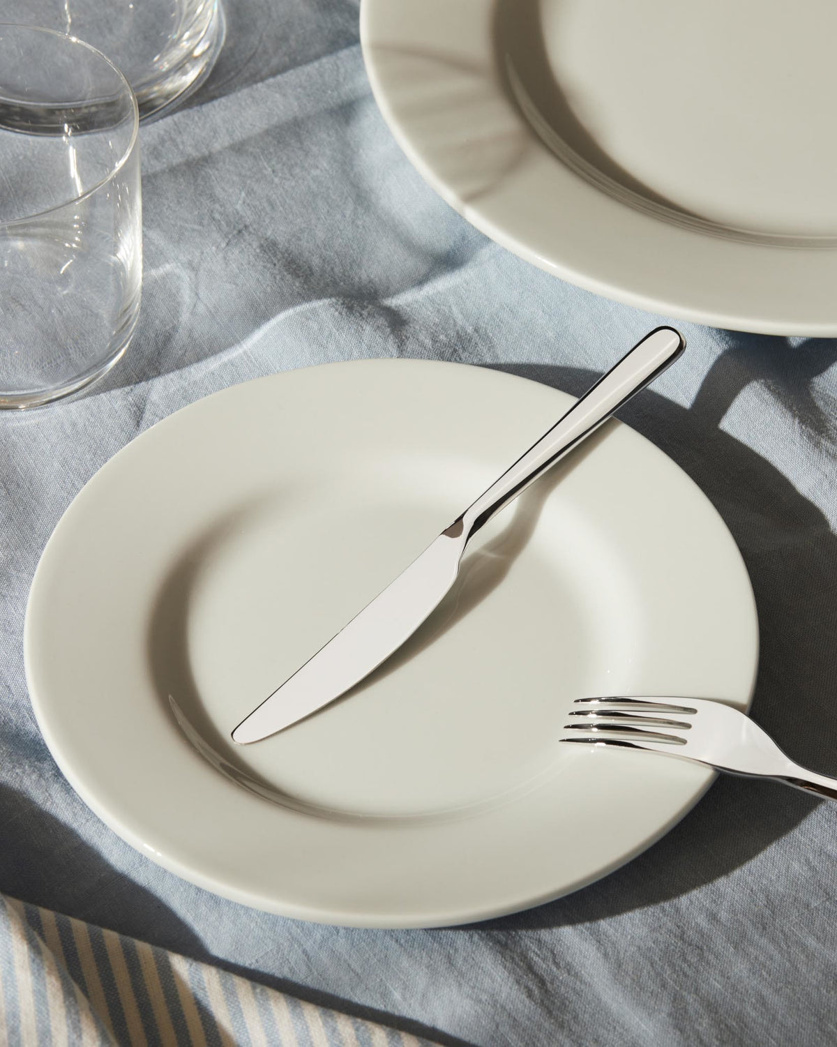 Shiny silver Amici cutlery set by Alessi, with a knife and fork on a white plate and light blue linen tablecloth.