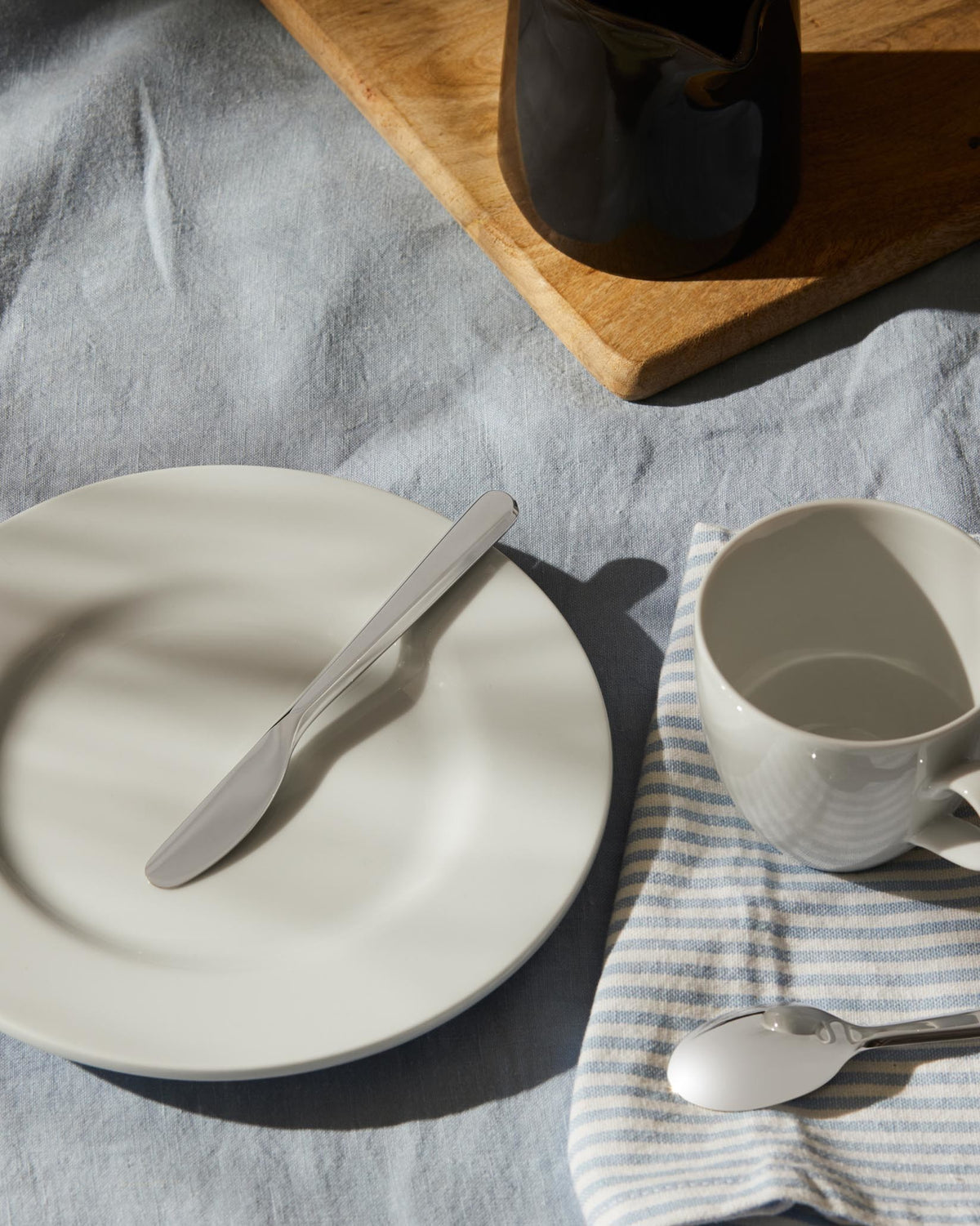 Sleek silver knife by Alessi resting on a white plate, with a blue and white striped napkin and white cup on a light blue tablecloth.