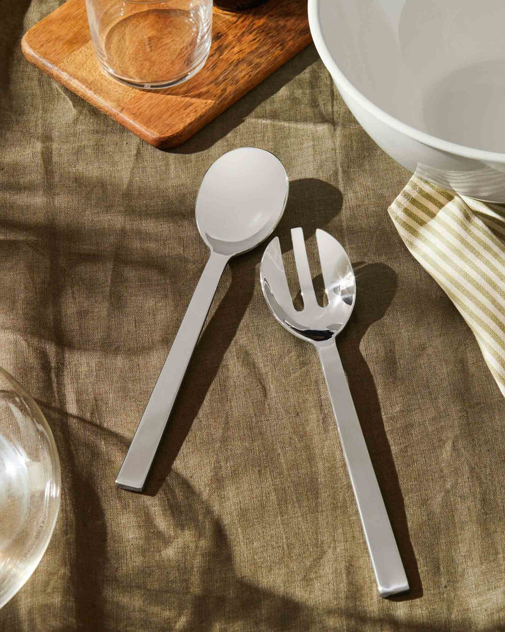 Shiny silver serving spoon and fork by Alessi, on a brown linen tablecloth next to a white bowl and striped napkin.