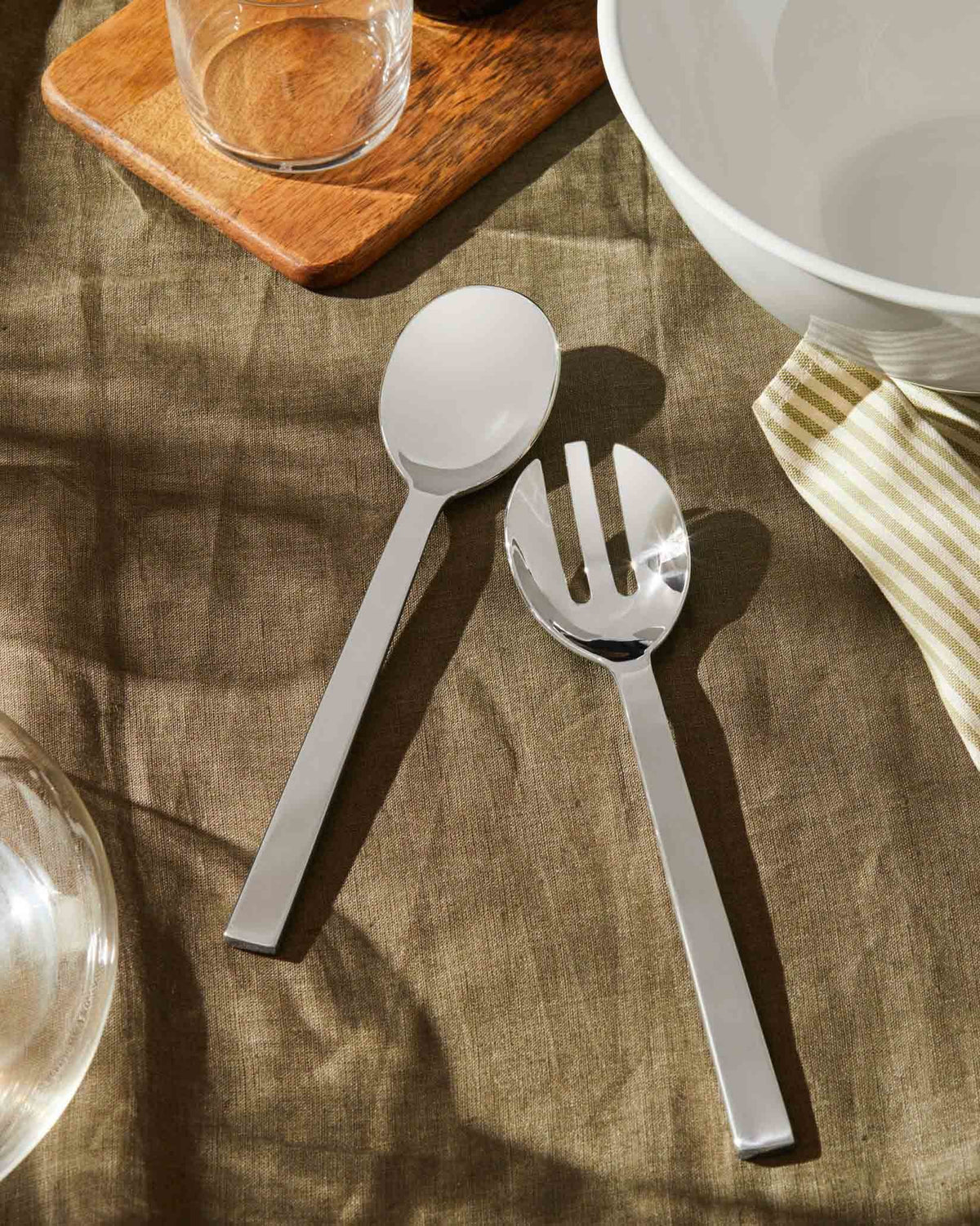 Shiny silver serving spoon and fork by Alessi, on a brown linen tablecloth next to a white bowl and striped napkin.