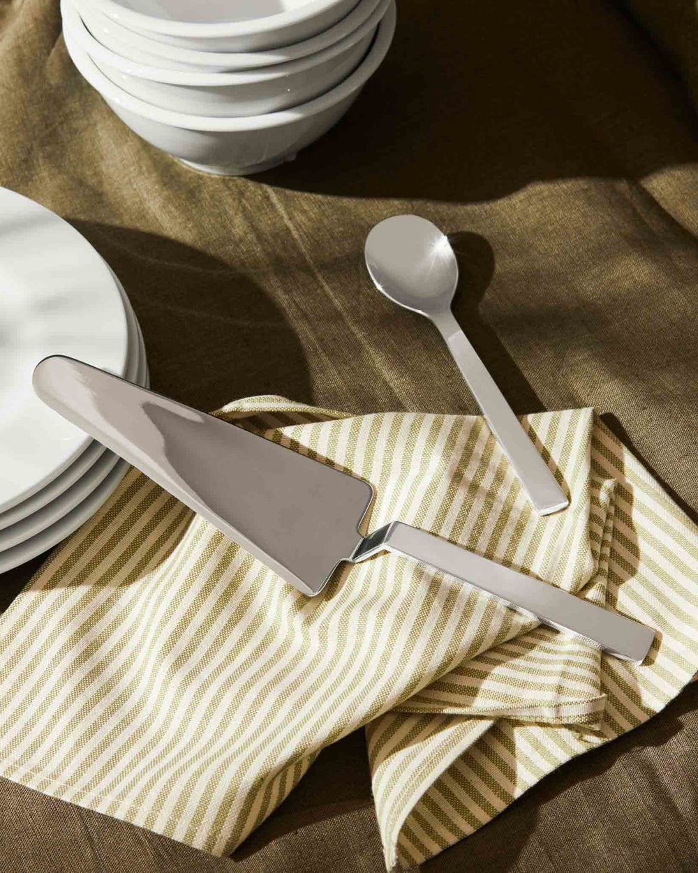 Silver cake server by Alessi, with a matching spoon on a striped green and white napkin, beside white plates.