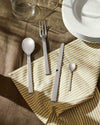 Polished stainless steel cutlery set by Alessi, arranged on a beige striped napkin with a brown tablecloth.
