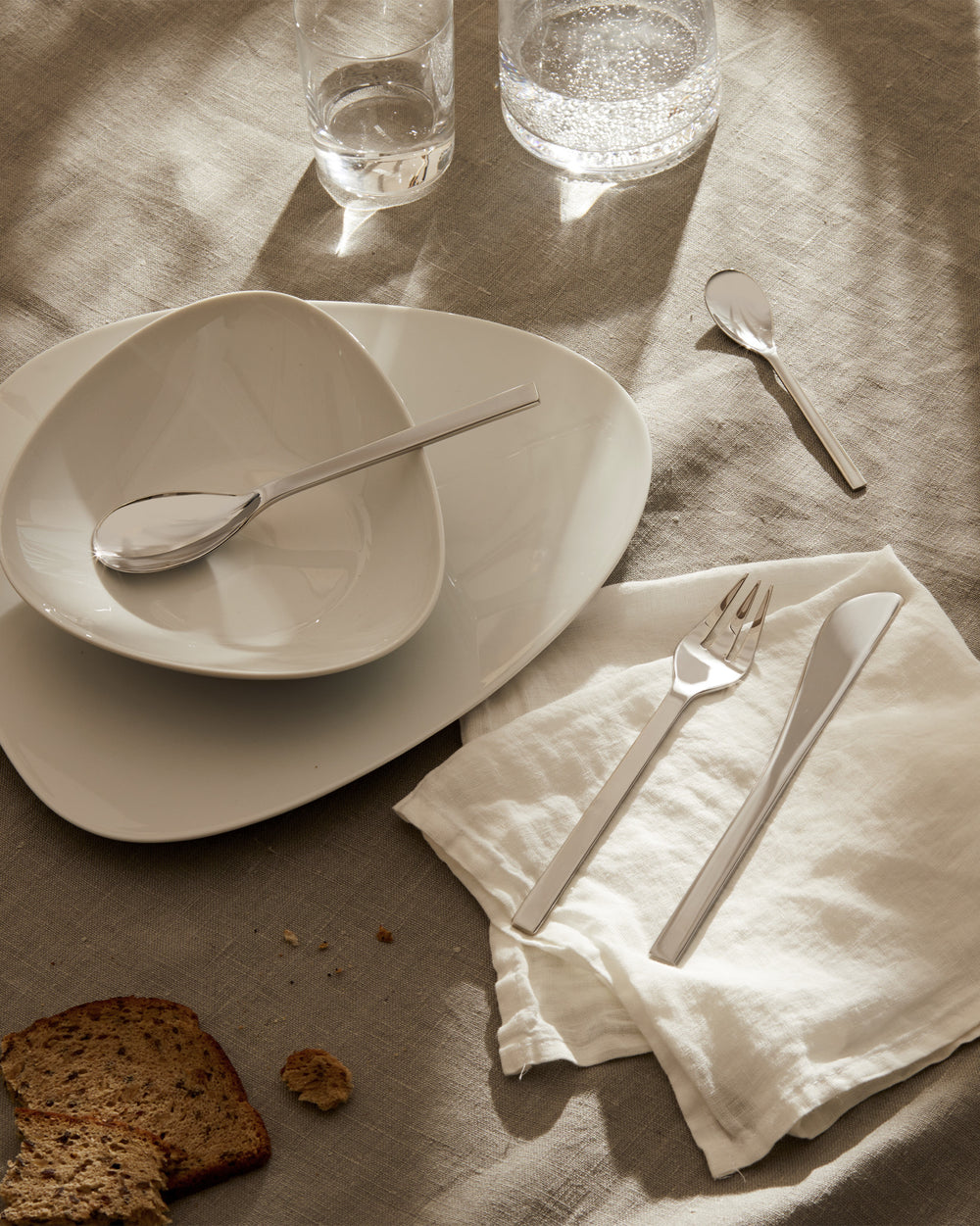 Colombina collection tableware by Alessi, including sleek stainless steel cutlery and white ceramic plates, on a beige linen tablecloth.