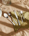 Silver stainless steel cutlery set by Alessi, including a knife, two forks, and two spoons, on a green linen napkin.