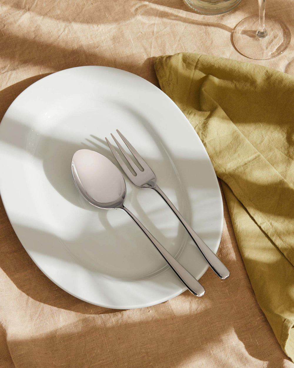 Silver stainless steel spoon and fork by Alessi on a white plate, with a beige linen tablecloth and green napkin.