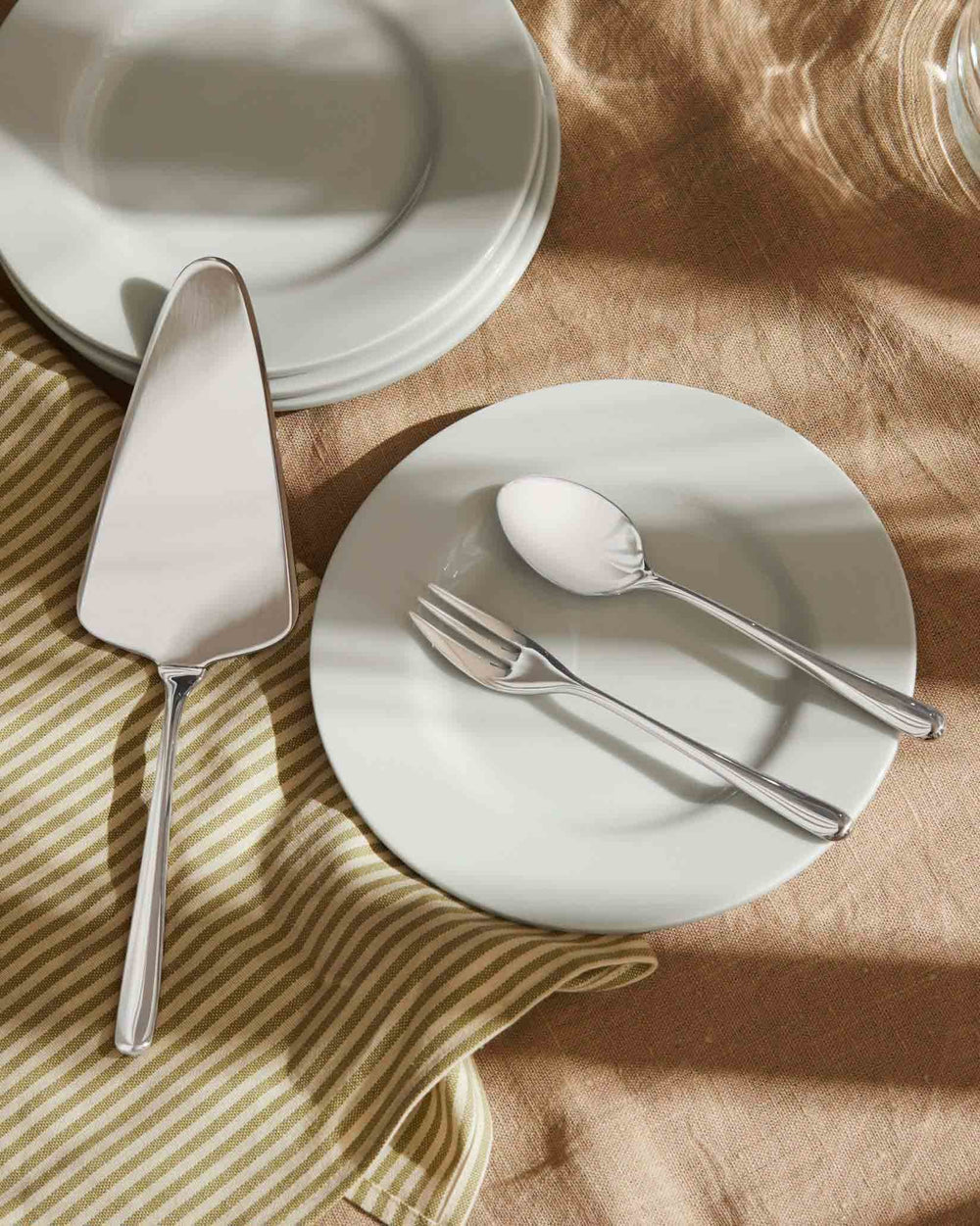 Silver cake server by Alessi, elegantly styled with matching cutlery on light gray plates and a striped napkin.