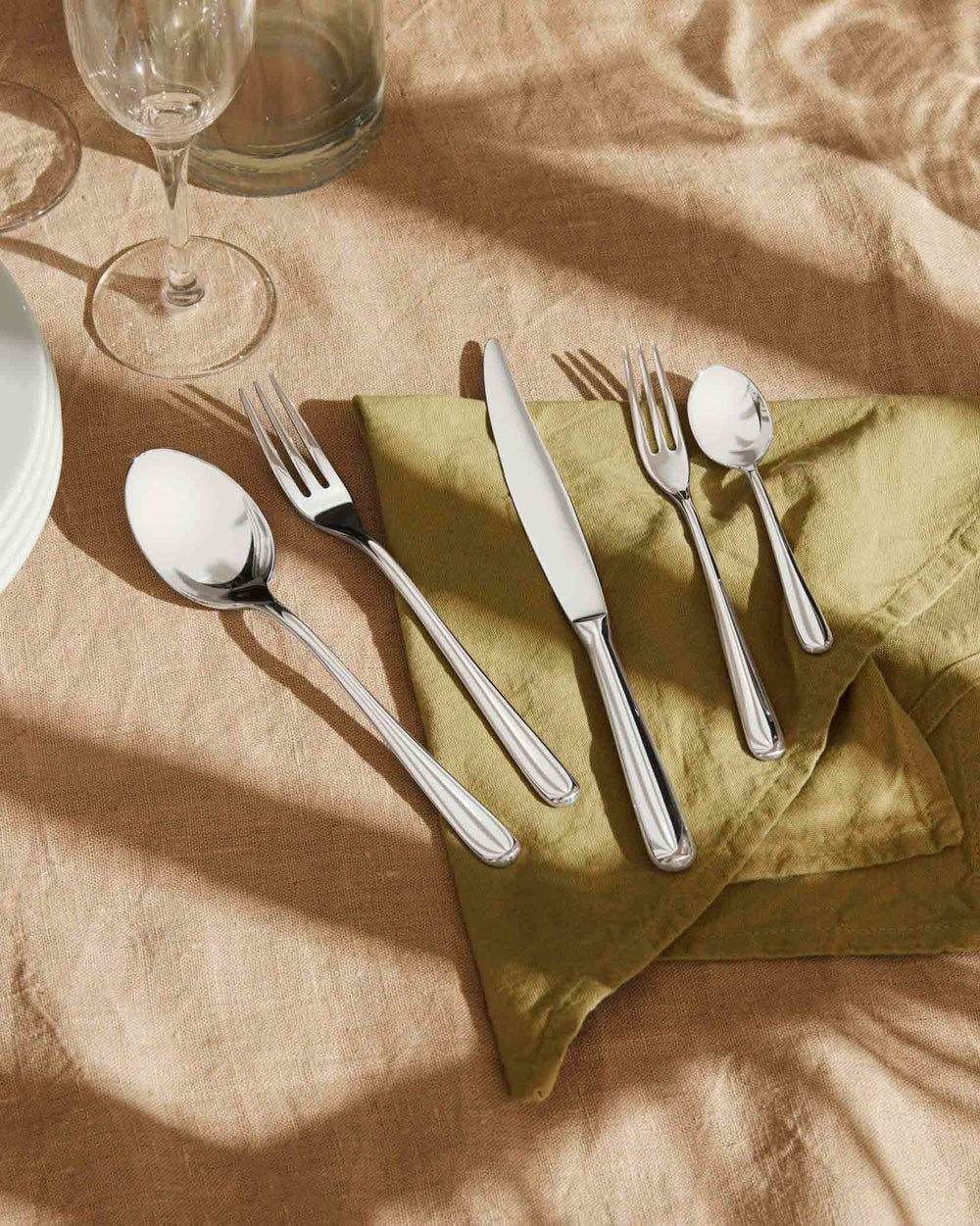 Sleek silver cutlery set by Alessi, displayed on a green napkin and light brown tablecloth.