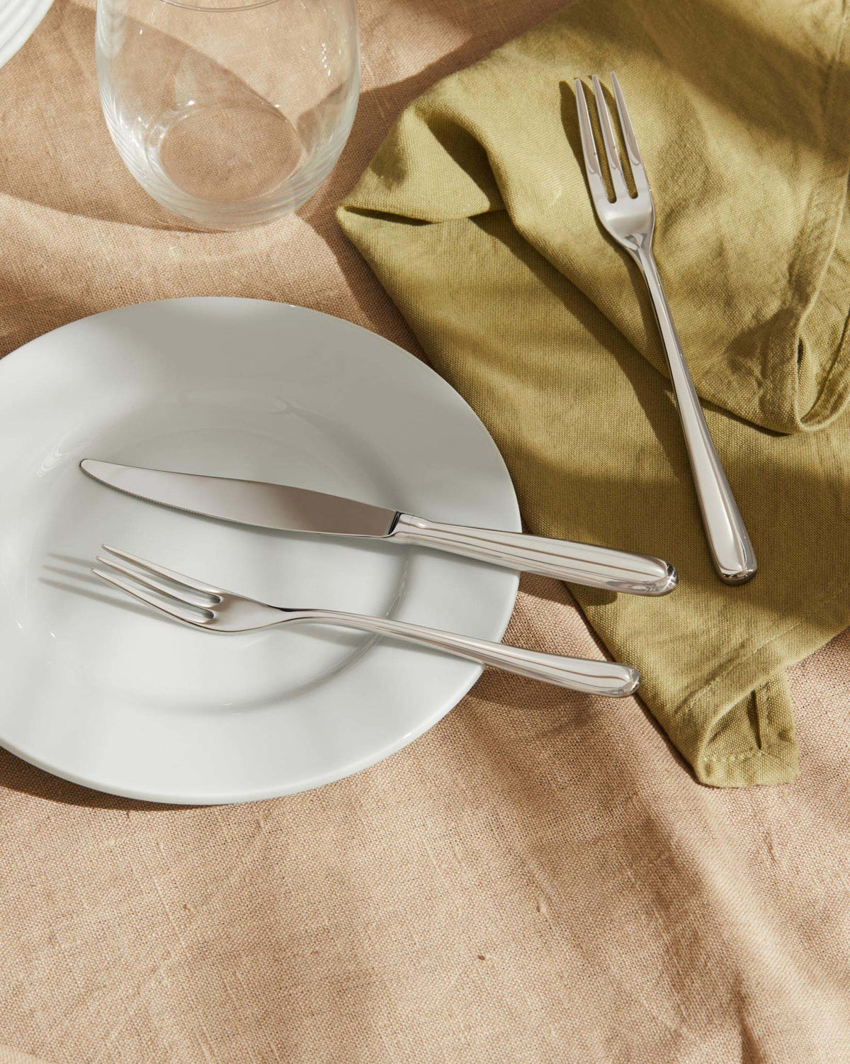 Sleek silver-toned cutlery set by Alessi on a white plate, green napkin, and linen tablecloth.