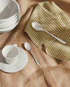Silver metallic spoons by Alessi resting on a striped green napkin and a brown textured tablecloth.