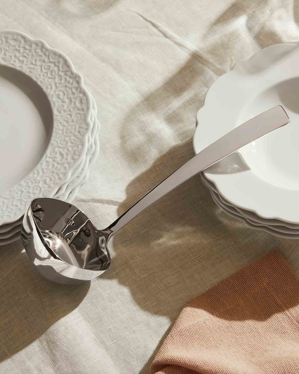 Polished silver serving ladle by Alessi, angled on a beige linen tablecloth with stacked white scalloped plates.