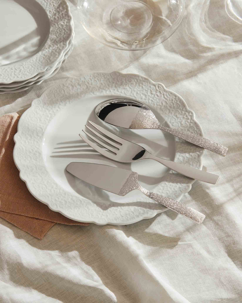 Silver cutlery pieces by Alessi with embossed ornate handles, on a white patterned plate.