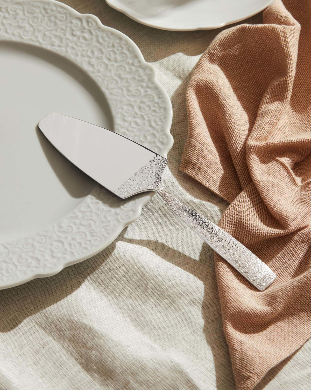 Silver cake server by Alessi with ornate embossed scrollwork handle, resting on a decorative white plate.