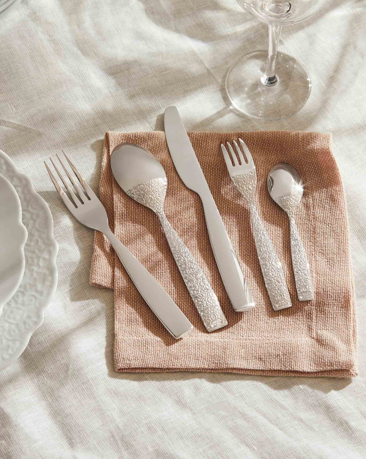 Silver stainless steel cutlery set by Alessi with embossed ornate handles, displayed on a pink napkin with plates.