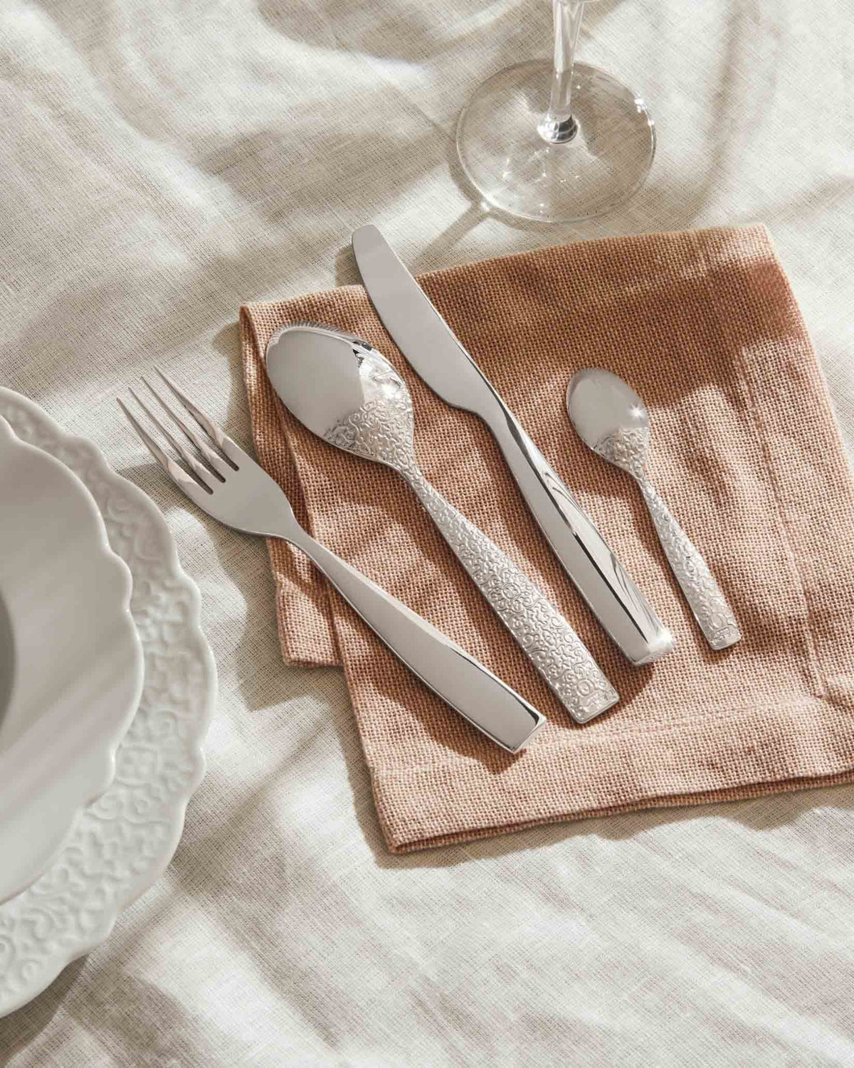 Silver flatware set with ornate embossed handles by Alessi, laid on a textured brown napkin beside white plates and a wine glass.