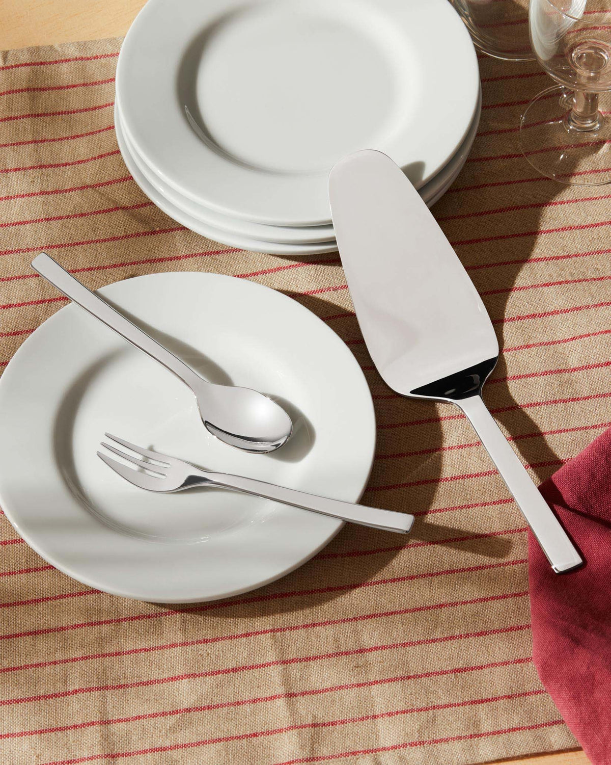 White Ovale plates and polished flatware by Alessi, including a spoon, fork, and cake server, on a red-striped runner.