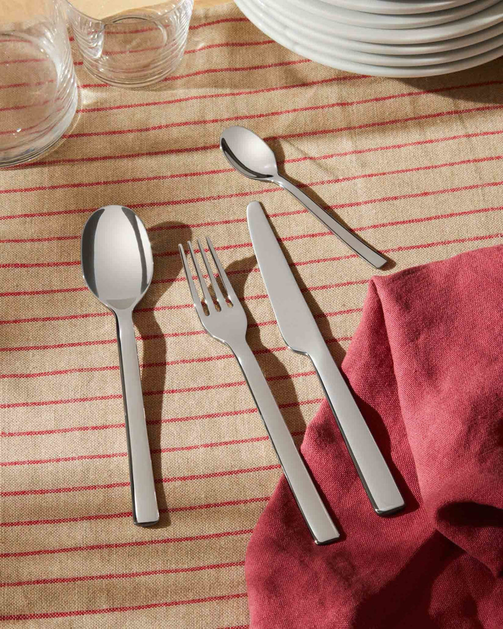 Stainless steel cutlery set by Alessi, including a spoon, fork, and knife, on a striped linen tablecloth.