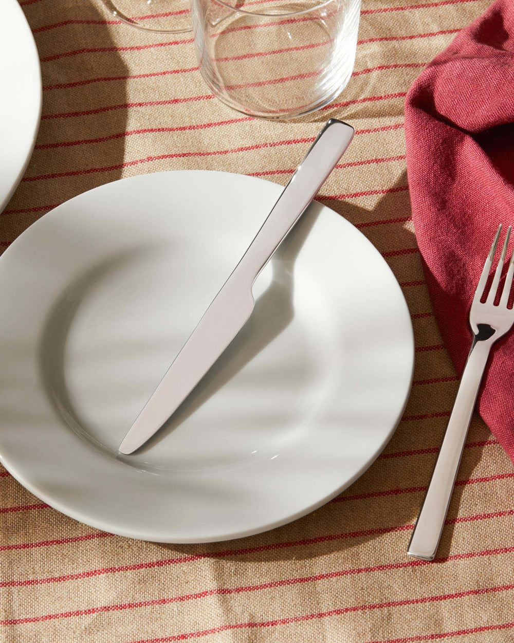 White Ovale dining plate by Alessi, with a sleek silver knife and fork on a striped tablecloth.