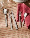 Four-piece silver stainless steel cutlery set by Alessi, displayed on a striped tablecloth with a red napkin.