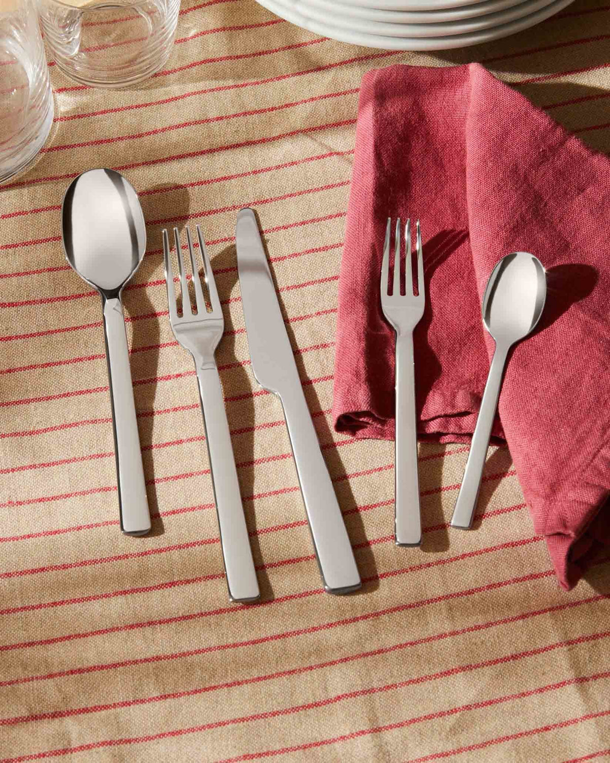 Stainless steel Ovale cutlery set by Alessi, elegantly laid on a beige striped tablecloth with a red napkin and white plates.