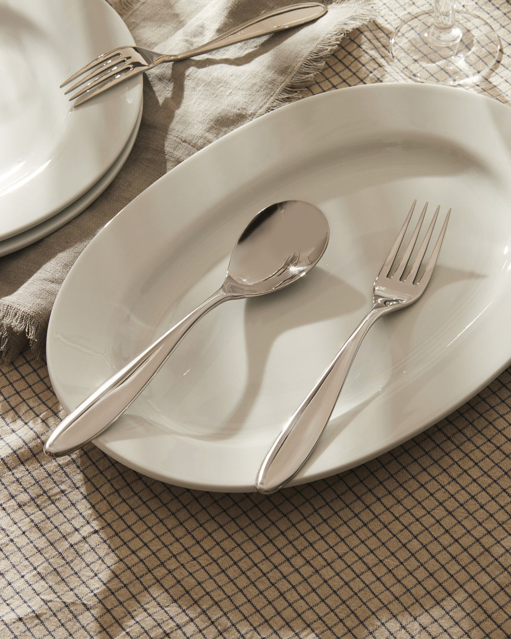 Sleek silver cutlery set by Alessi, featuring a spoon and fork on a white oval plate, atop a checkered tablecloth.