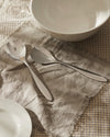 Silver Mami salad servers by Alessi, resting on a fringed linen napkin over a checkered tablecloth.