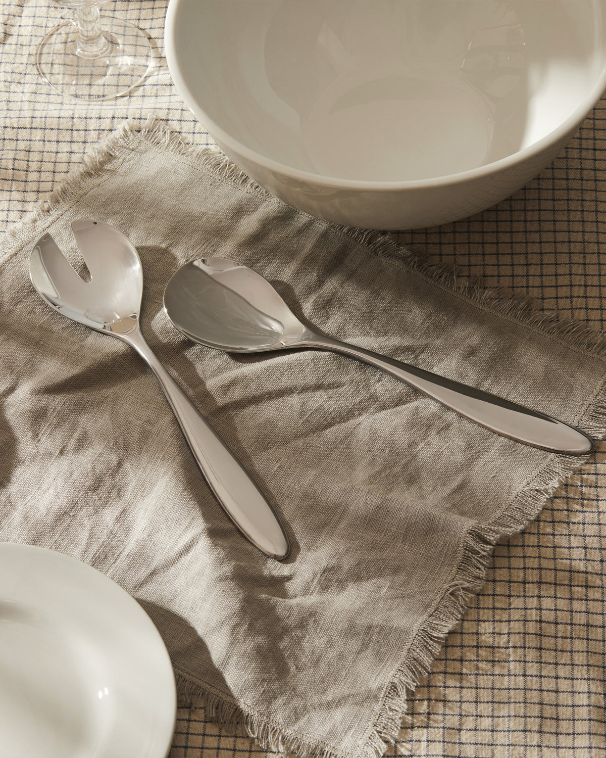 Silver Mami salad servers by Alessi, resting on a fringed linen napkin over a checkered tablecloth.