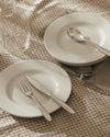 White ceramic tableware set by Alessi, with silver cutlery and clear glasses on a beige checkered tablecloth.