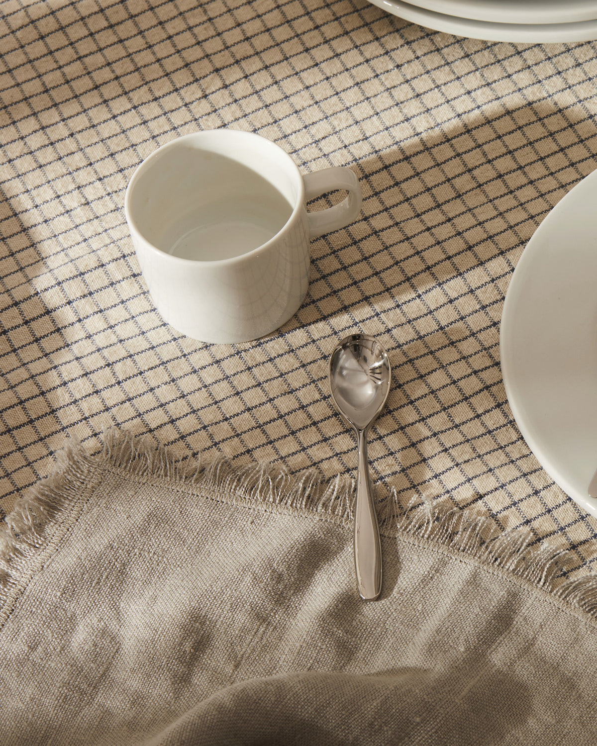 White ceramic Mami mug by Alessi, on a textured beige checkered tablecloth, with a silver spoon and white plate.