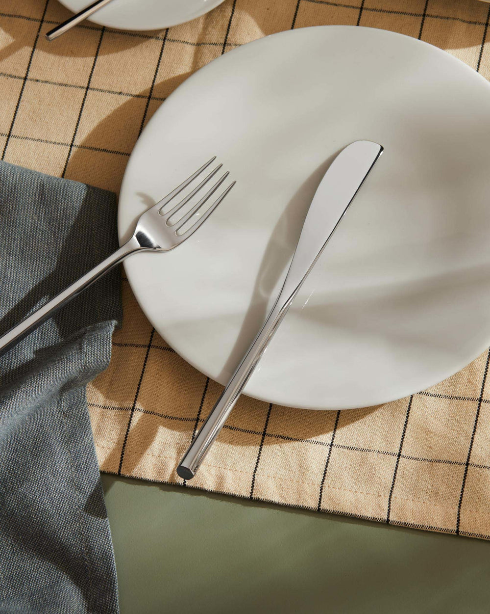 Stainless steel fork and knife by Alessi, sleek and modern, on a beige plaid placemat with a blue napkin.