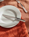 Silver dinner knife by Alessi, resting on a white plate with a fork on an orange linen tablecloth.