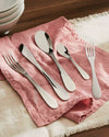 Sleek silver stainless steel flatware set by Alessi, arranged on a pink linen napkin and beige placemat.