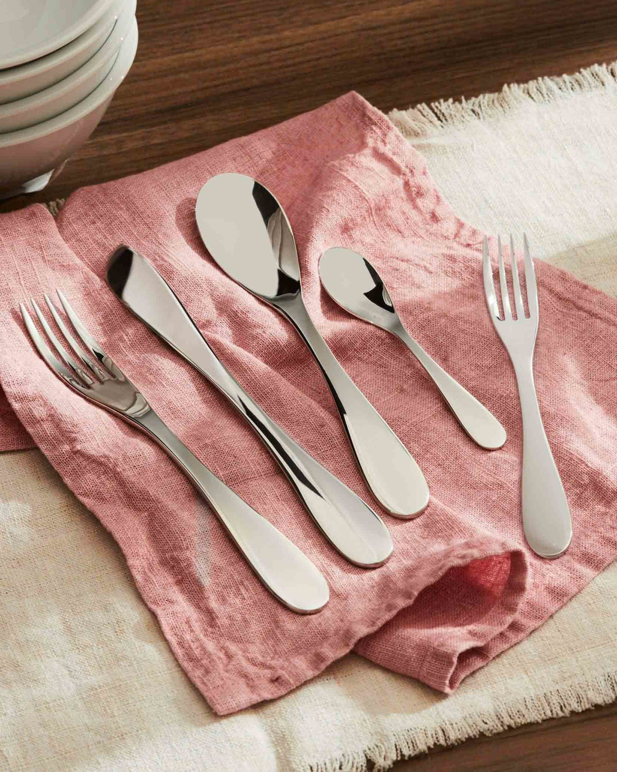 Sleek silver stainless steel flatware set by Alessi, arranged on a pink linen napkin and beige placemat.