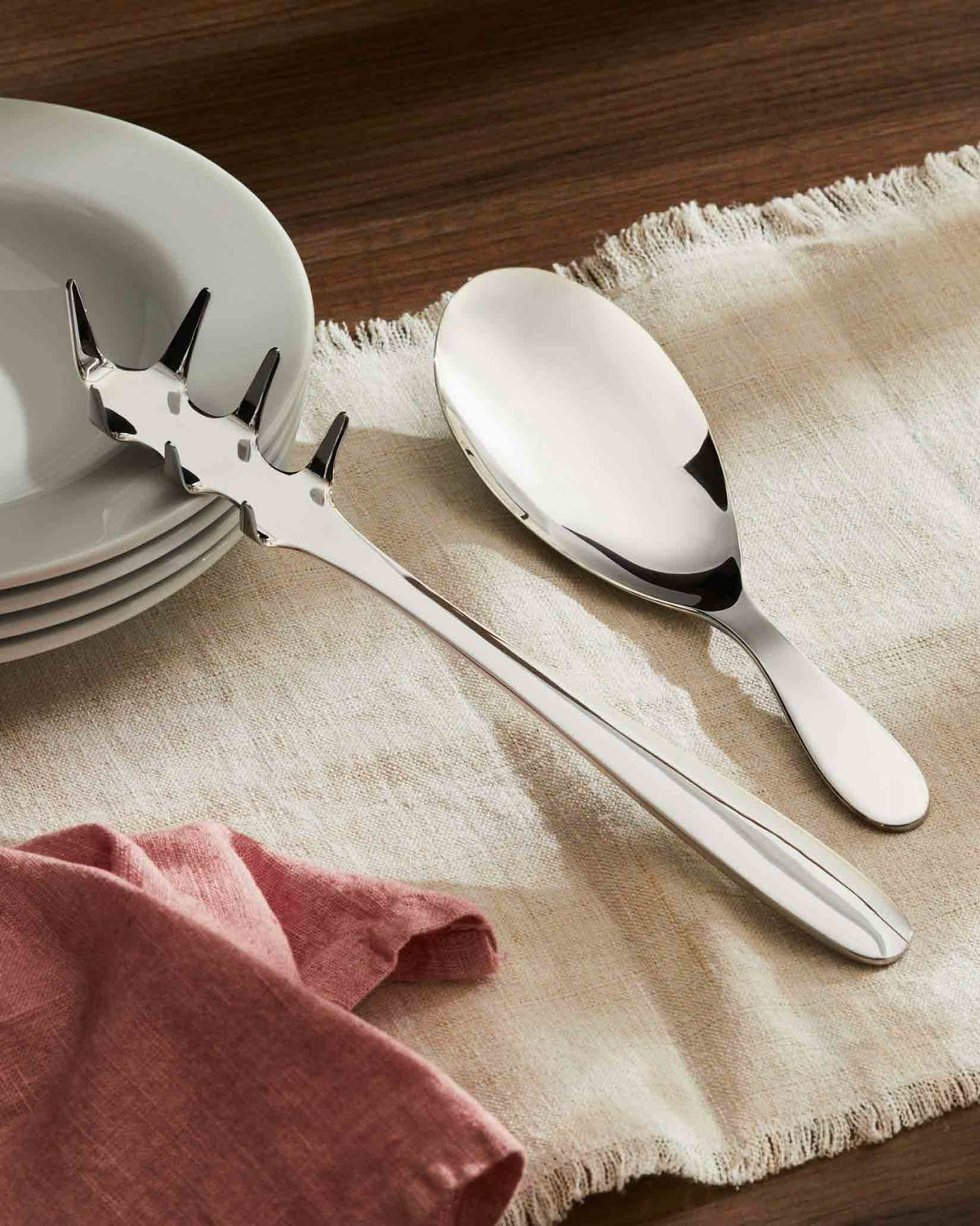 Sleek stainless steel serving utensils by Alessi, a pasta server and spoon on a fringed beige tablecloth.