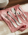 Silver stainless steel cutlery set by Alessi, elegantly laid on a pink linen napkin next to stacked white bowls.