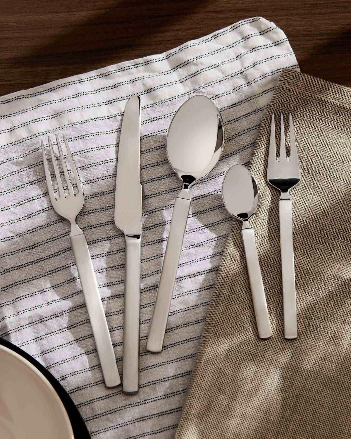 Dry silver-toned cutlery set by Alessi with sleek, flat handles on striped and plain linen napkins.