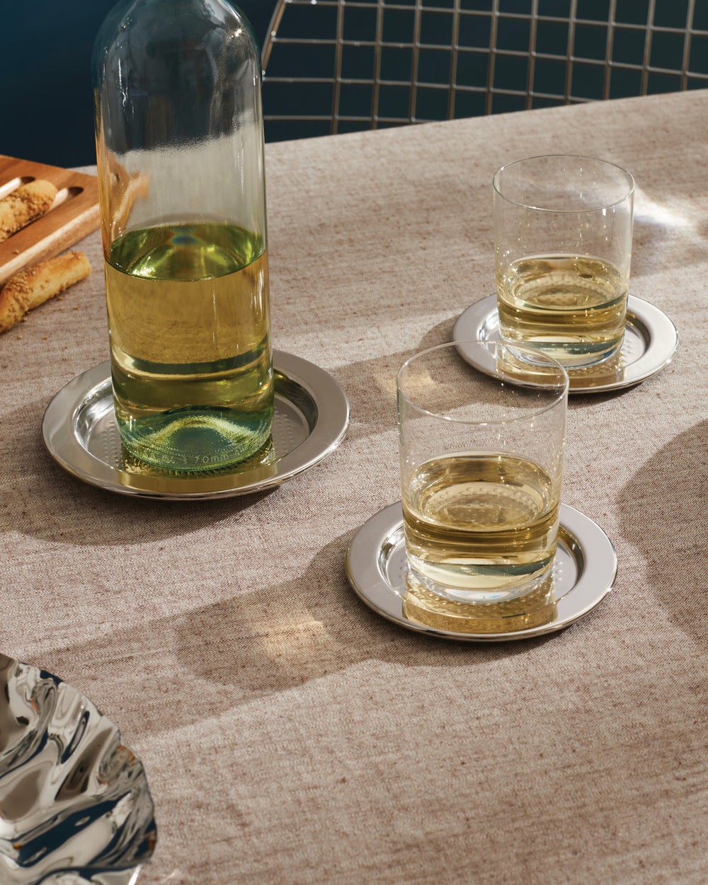 Silver metallic coaster with a dimpled surface by Alessi, under a clear glass bottle and two tumblers with light liquid on a linen tablecloth.