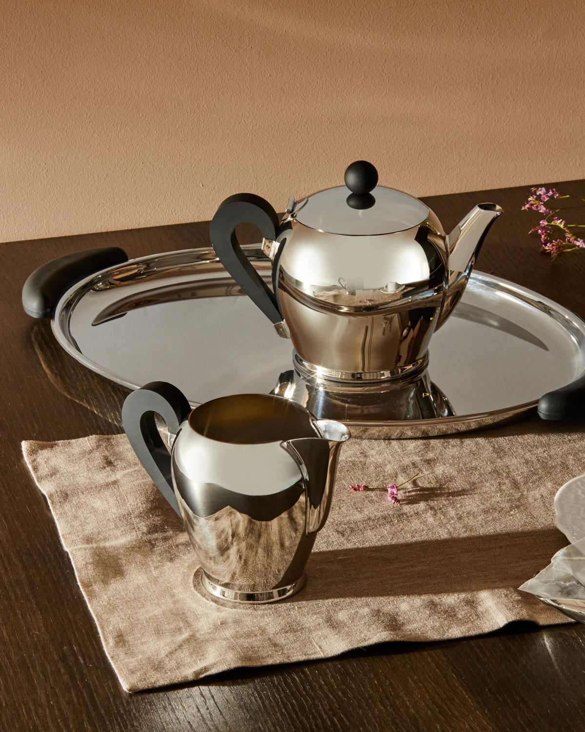 Shiny silver Bombé teapot by Alessi with black handles, a matching creamer, and a silver tray on a brown linen cloth.