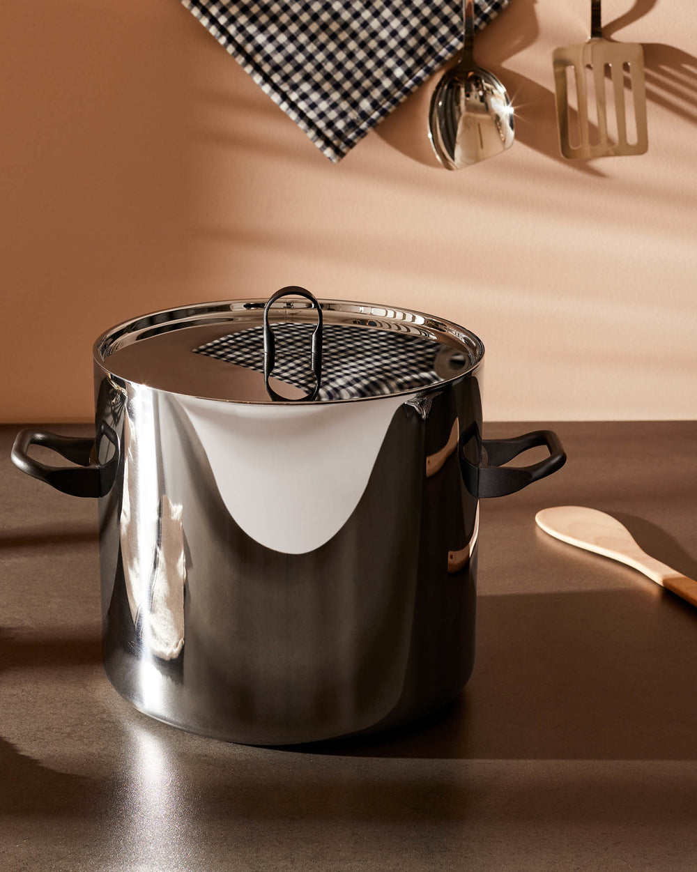 Shiny stainless steel stockpot by Alessi with black handles and a reflective lid showing a checkered cloth, on a dark countertop.