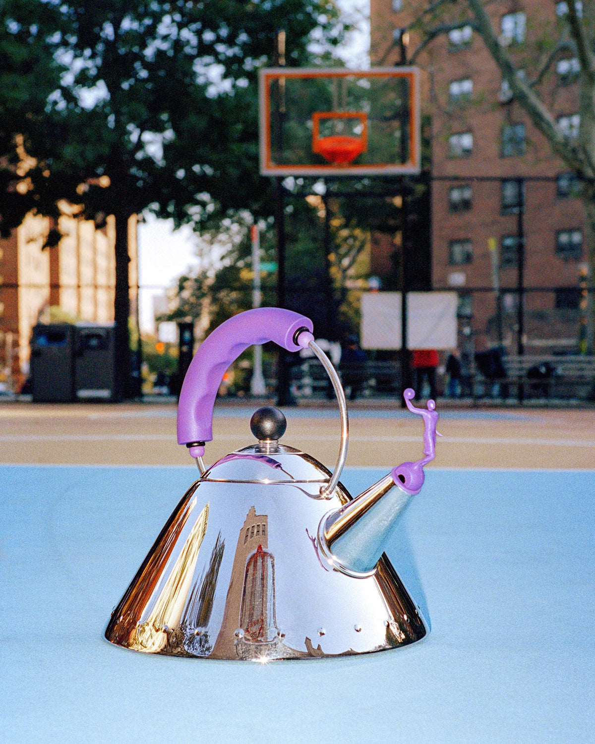 Reflective chrome conical kettle by Alessi with a purple handle and person-shaped whistle, on a blue court.