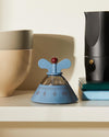 Light blue and chrome Kitchen Timer by Alessi with a red knob and wing-shaped handles, on a shelf with ceramic bowls and a moka pot.