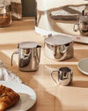 Polished stainless steel milk jug with a hinged lid and integrated handle by Alessi, beside a matching sugar bowl and small creamer on a wooden table.
