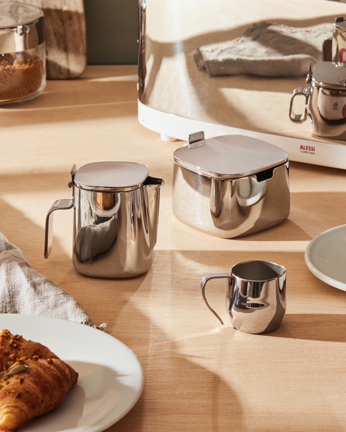 Silver-toned stainless steel creamer with a hinged lid by Alessi, displayed alongside a matching square sugar bowl and a small creamer cup on a wooden table.