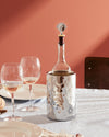 Silver metal wine stopper by Alessi with a whimsical face design, on a bottle in a patterned wine cooler.