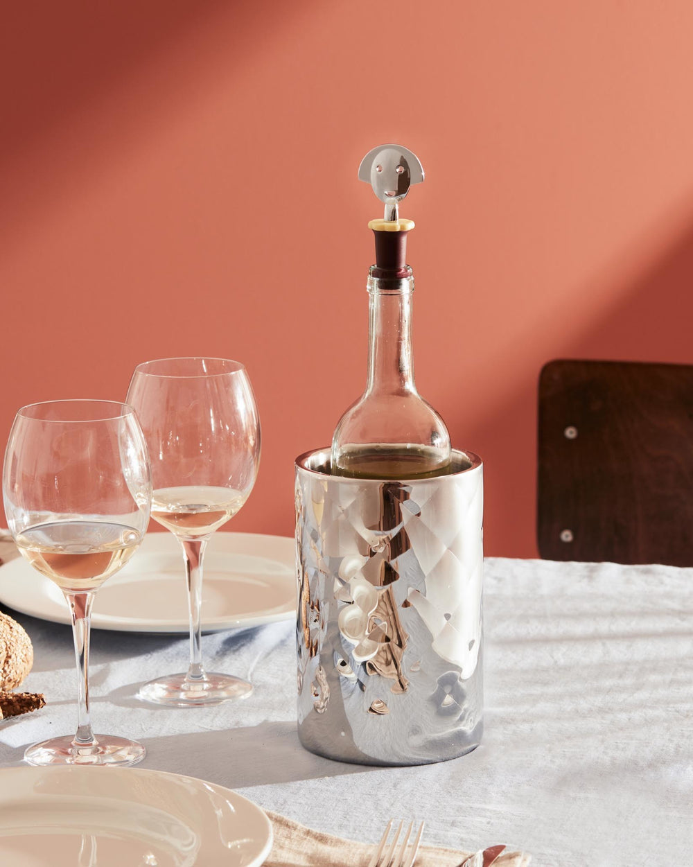 Silver metal wine stopper by Alessi with a whimsical face design, on a bottle in a patterned wine cooler.